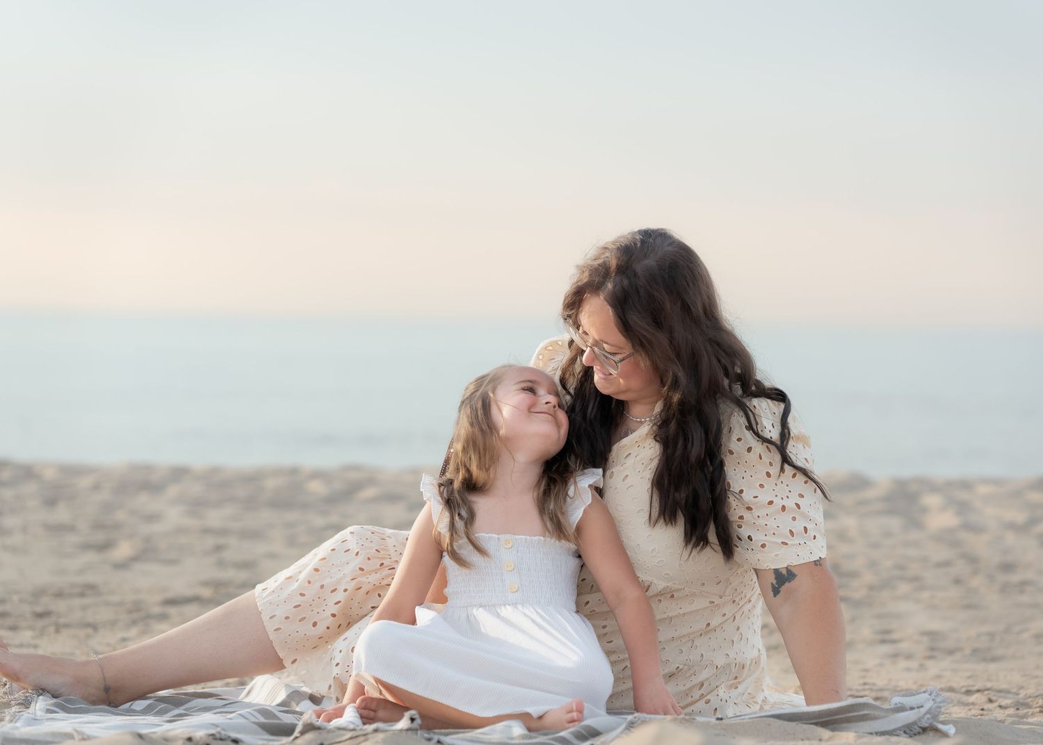 A mother and daughter share a sweet moment sitting on a blanket at the beach in matching white outfits.