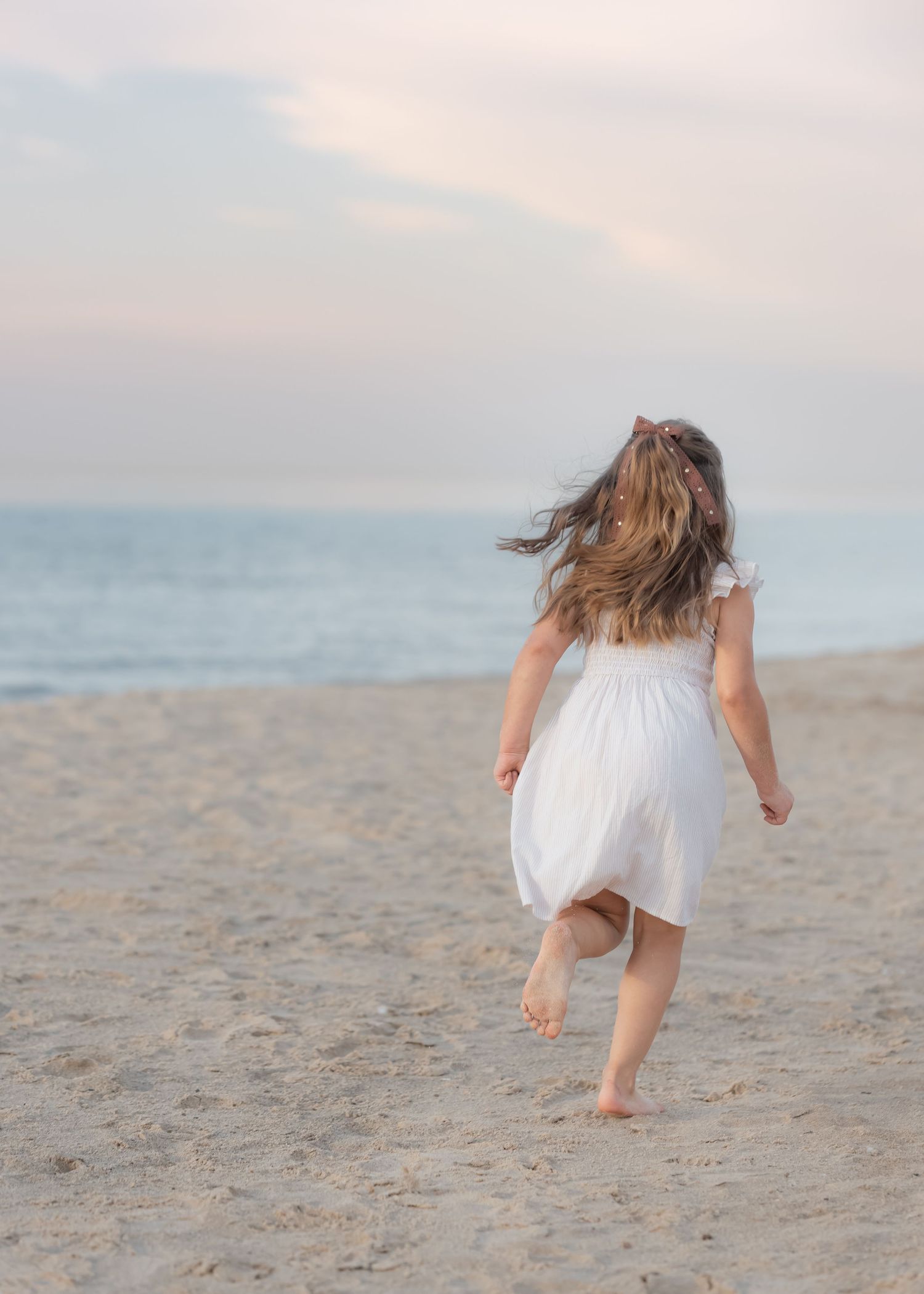 A young girl in a white dress runs barefoot on a sandy beach toward the ocean at sunset.