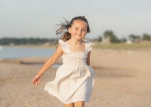 Young girl in white dress running joyfully on sandy beach, hair blowing in wind, with water and sky in background.