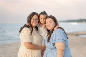 Three women smiling together on a beach at sunset, wearing pastel dresses in a warm family portrait.