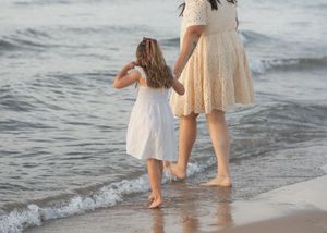 A mother and young daughter walk hand-in-hand along a sandy beach shore in matching white dresses.