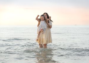 A mother carries her laughing child on her back while wading in ocean waves at sunset on a beach.