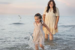 A young girl splashes in ocean waves while a woman in a yellow dress watches at sunset on a sandy beach.