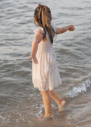 A young girl in a white dress plays at the ocean shoreline, her long hair blowing as waves wash over her bare feet.