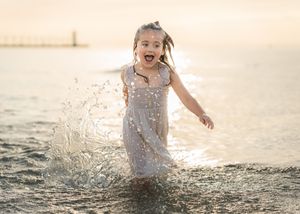 Young girl in white dress splashing joyfully in ocean waves at golden sunset near lighthouse.