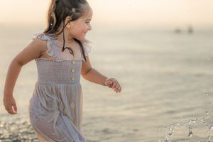 A young girl in a gray sundress runs joyfully along a sandy beach at sunset, splashing water.