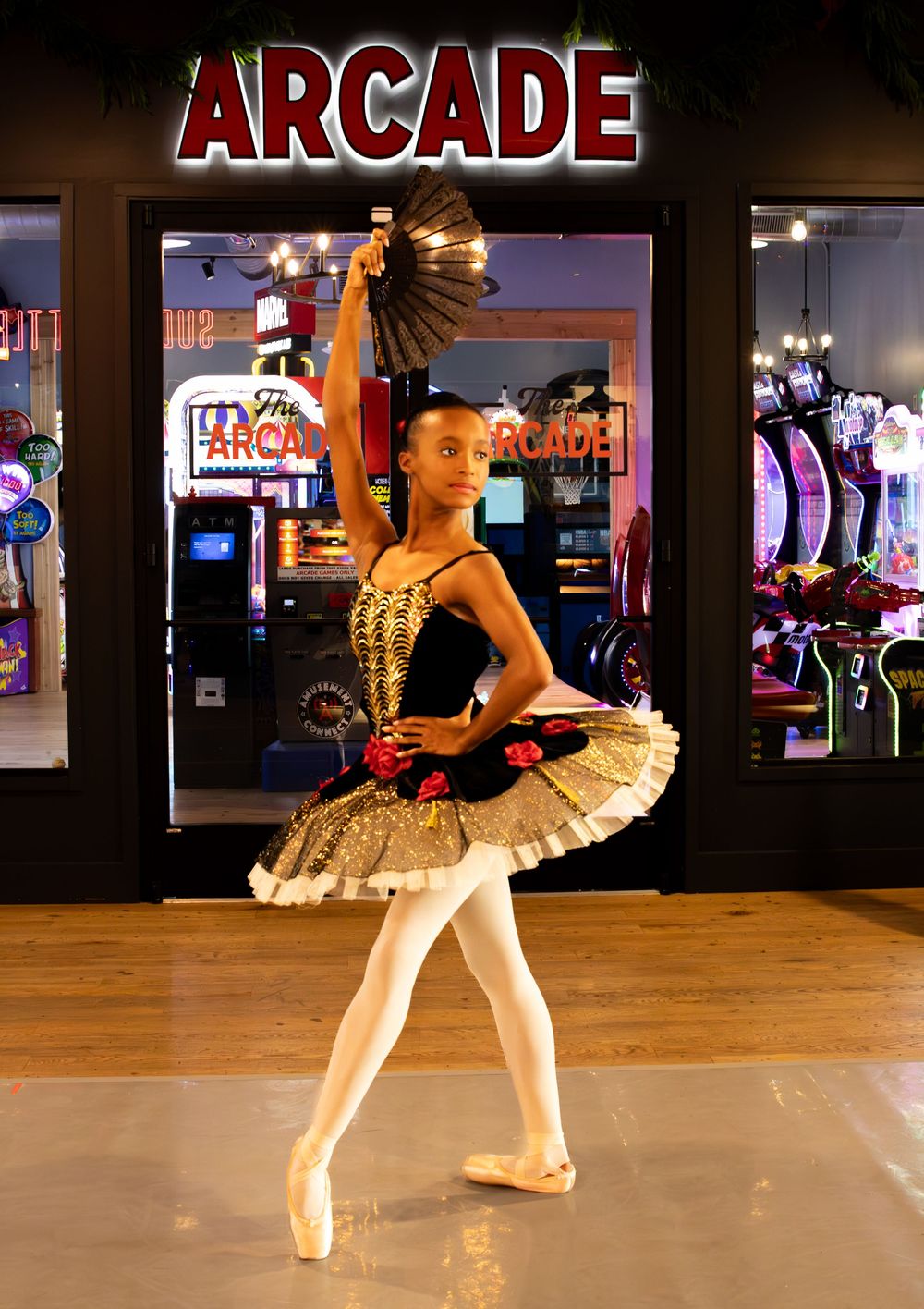 Young ballerina in gold costume on pointe holding black fan outside arcade, performing classical ballet.