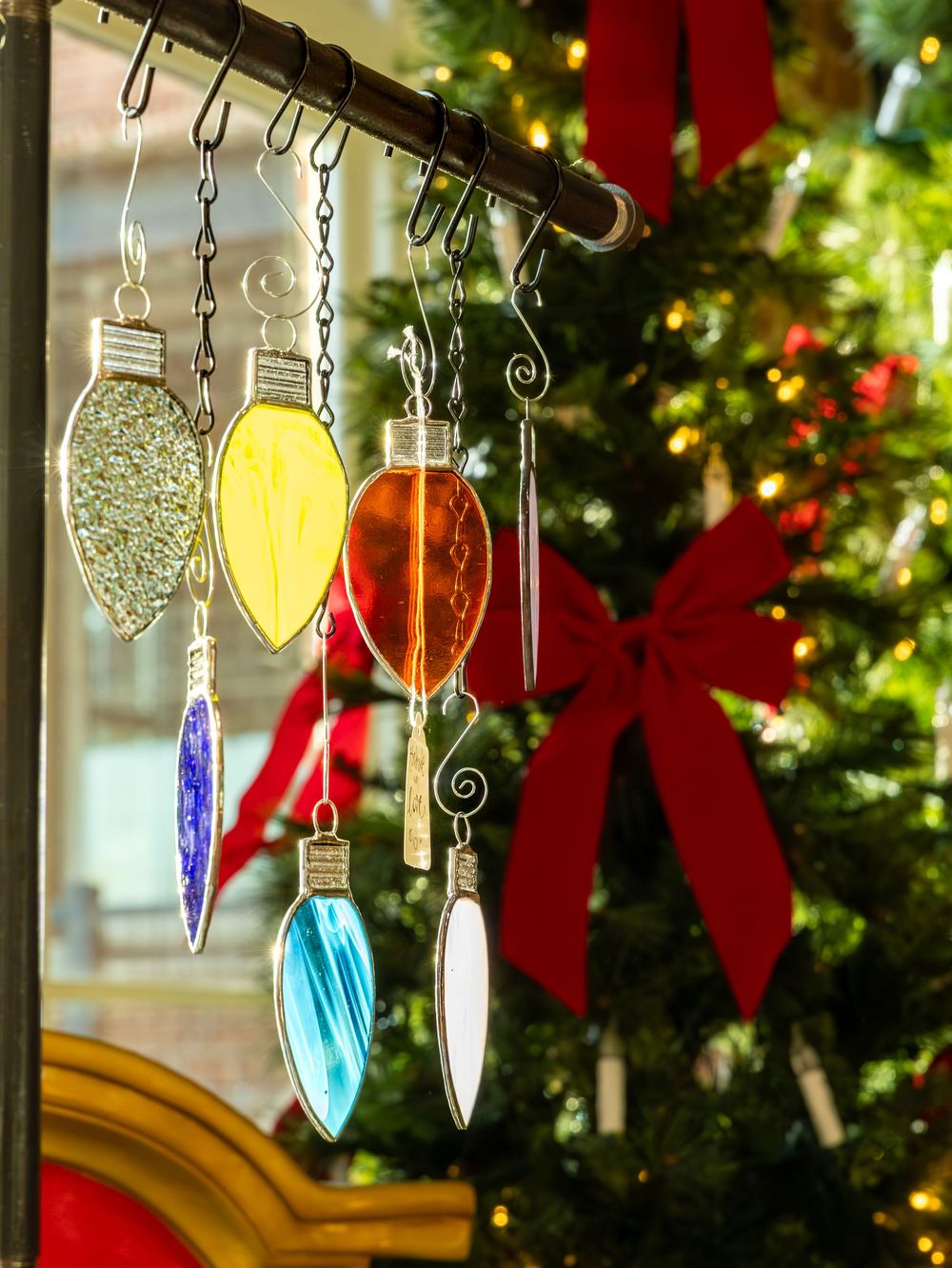 Colorful stained glass Christmas light ornaments hanging from a rod with a decorated tree in the background.