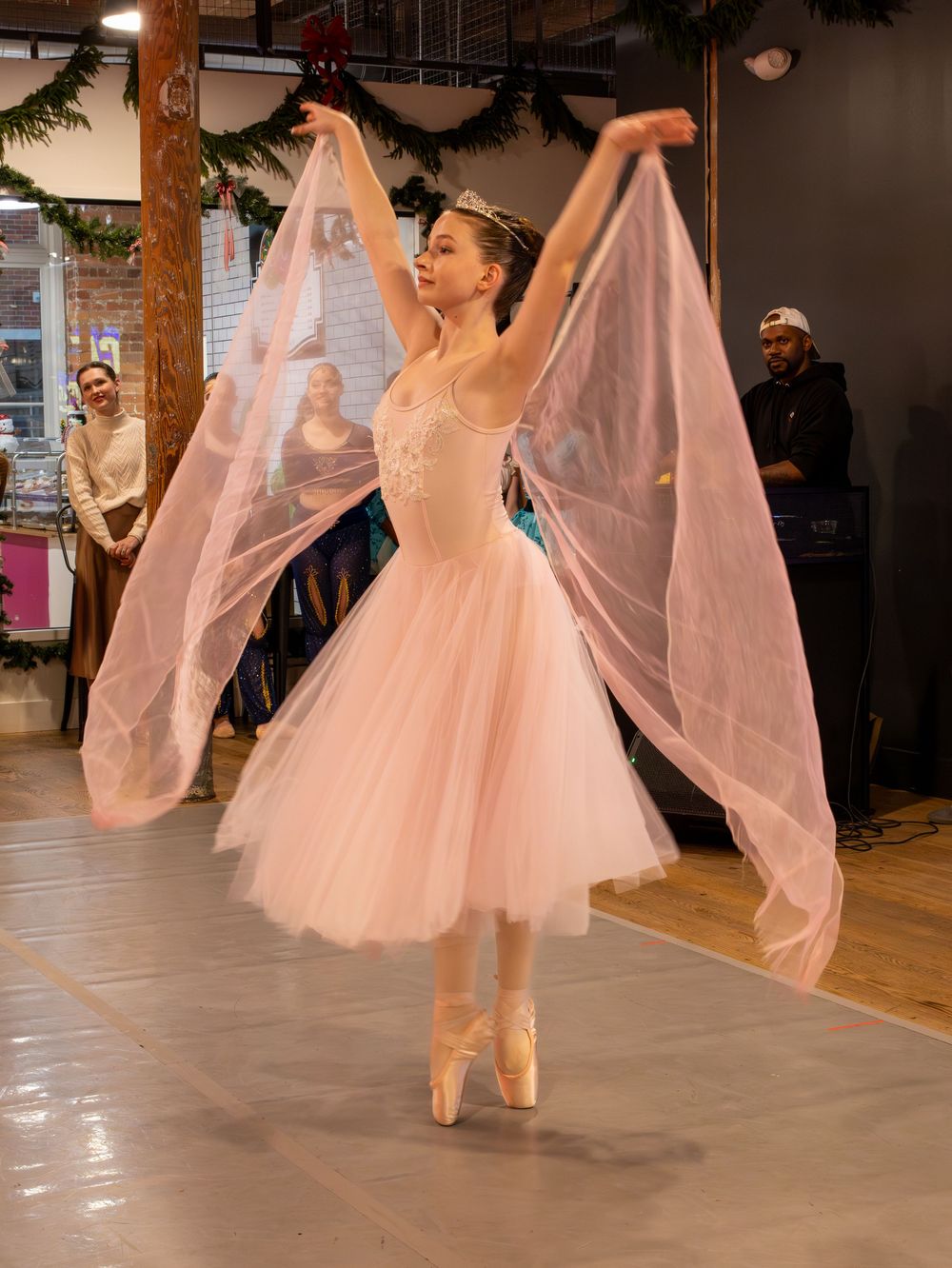 A ballerina in a pink tutu performs on pointe with flowing fabric wings in an indoor holiday setting.