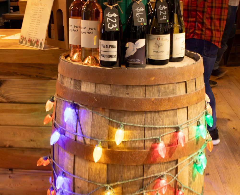 Wine bottles displayed on a rustic wooden barrel wrapped with colorful Christmas string lights.