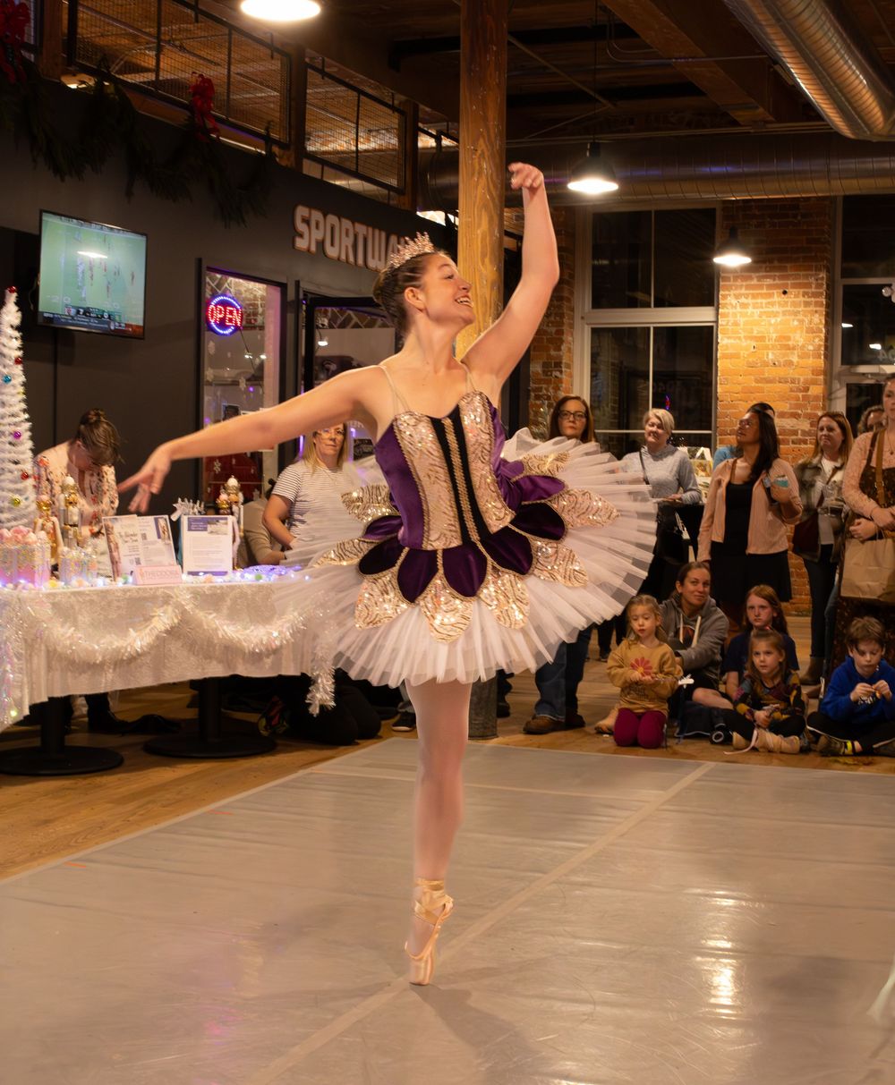 A ballerina in a purple and gold tutu performs on pointe before a crowd in an indoor venue.