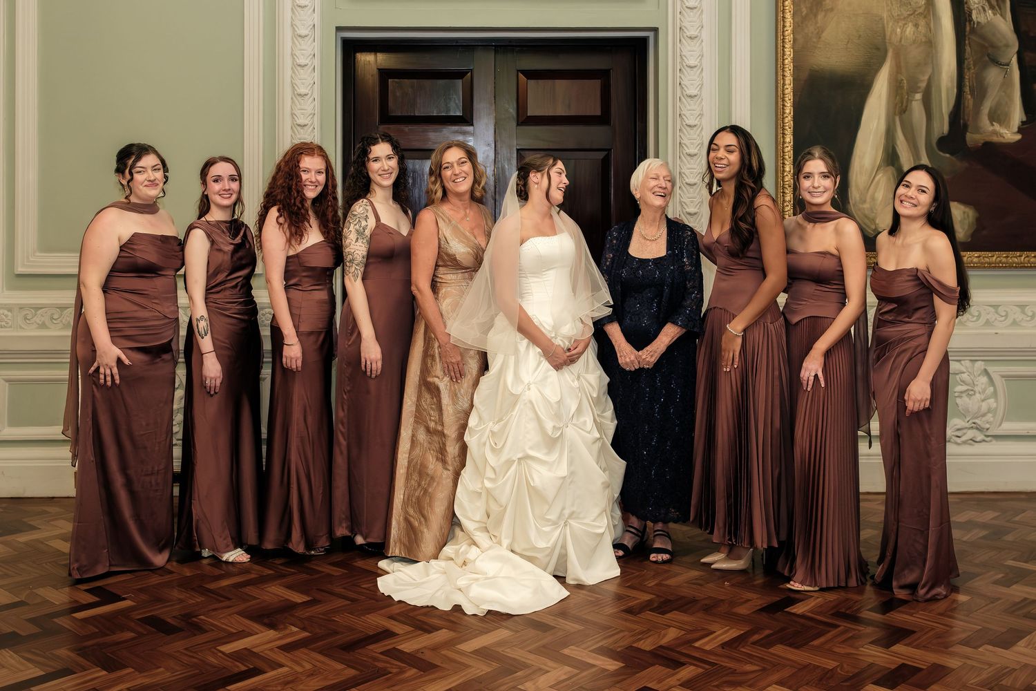 Bride in a white ball gown poses with bridesmaids in chocolate brown dresses and an older woman in navy in an elegant paneled room.