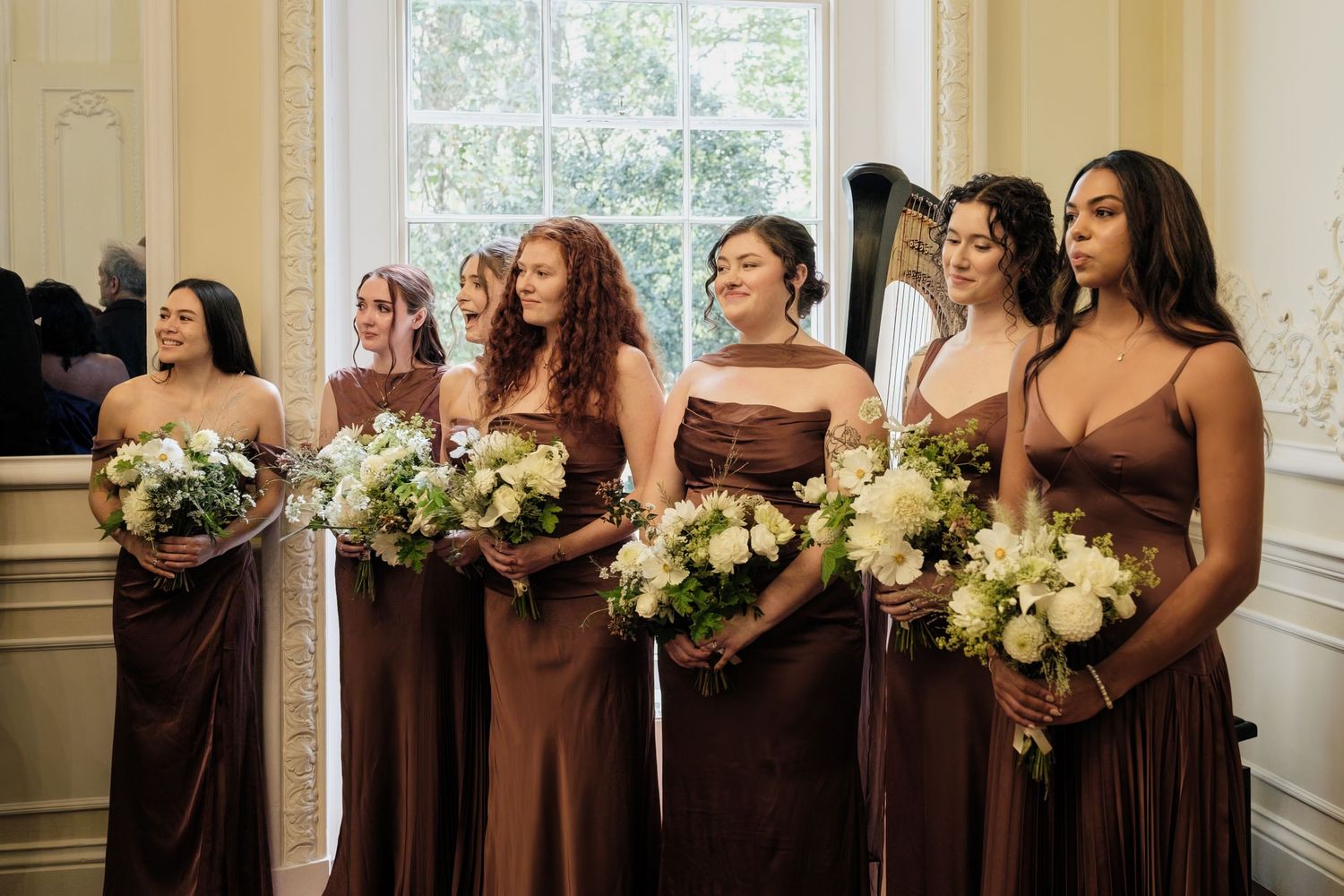 Six bridesmaids in brown satin dresses hold white floral bouquets while standing together in a bright room with tall windows.