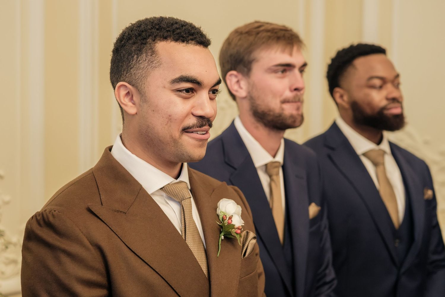 Three groomsmen in a row at a wedding, the man in front wearing a brown suit with a white boutonniere, flanked by two men in navy suits.