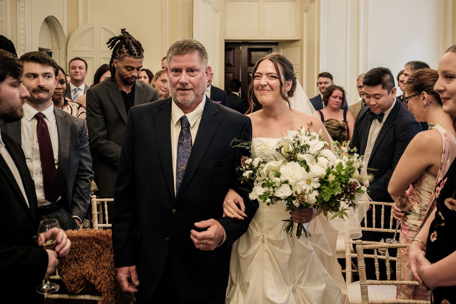 Bride in a white gown holding a white bouquet walks down the aisle on the arm of a smiling man in a dark suit as wedding guests look on.