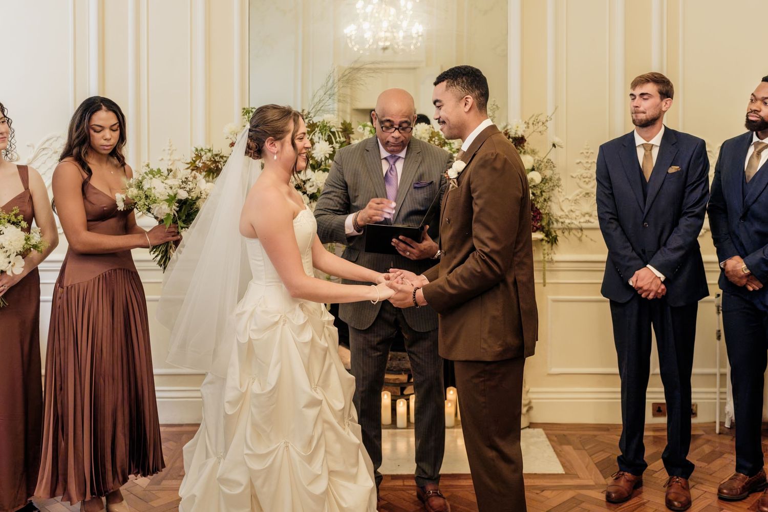 Bride in white gown and groom in brown suit exchange vows during an indoor wedding ceremony as an officiant and bridal party look on.