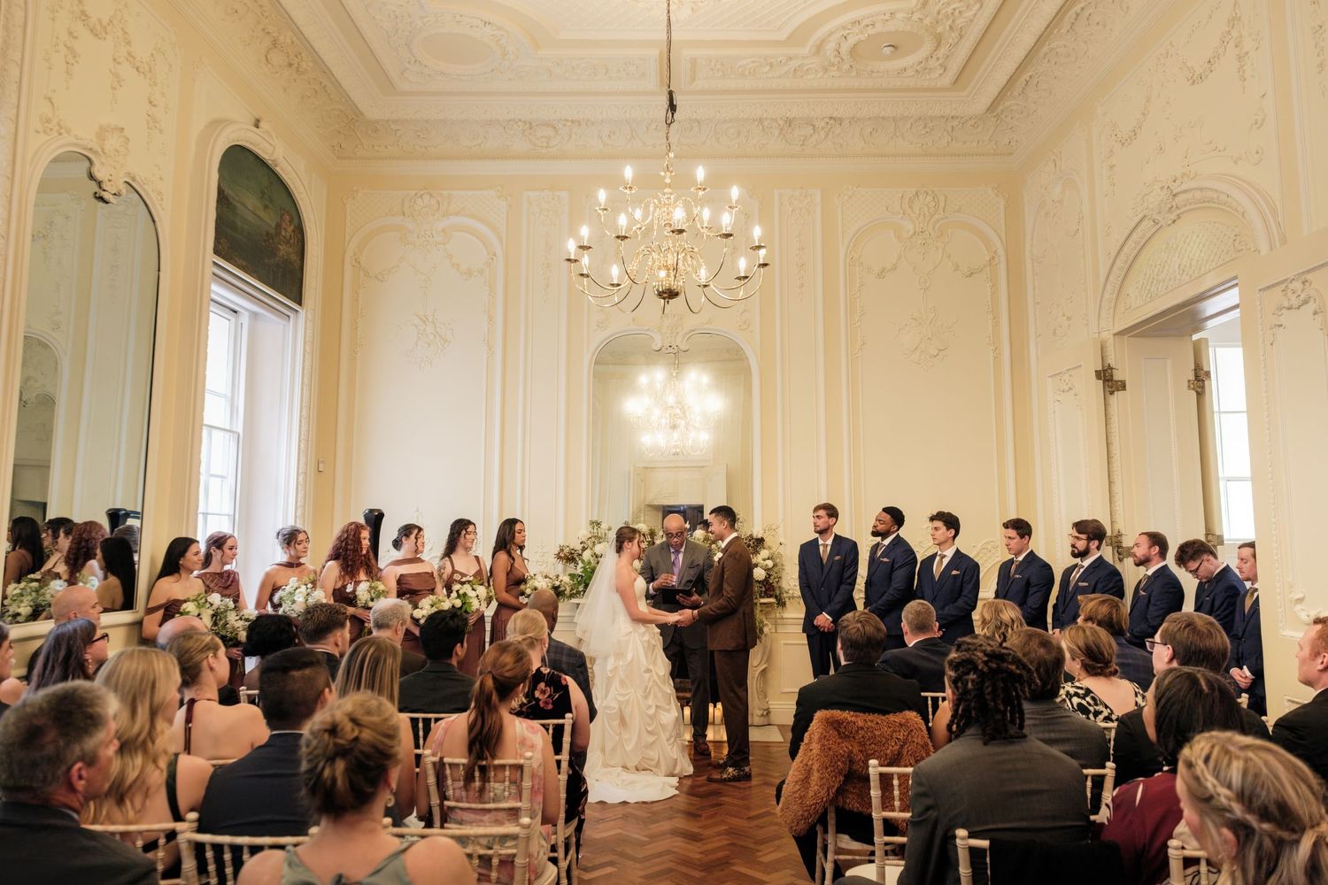 Bride and groom exchange vows at an elegant indoor wedding ceremony with groomsmen, bridesmaids, and guests in a grand ballroom with chandel