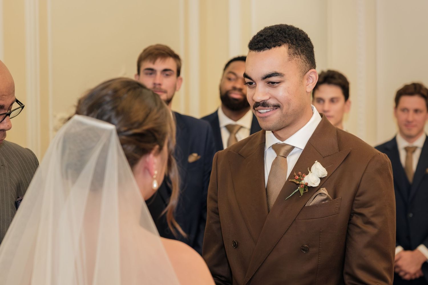 Smiling groom in a brown suit with a boutonniere faces his veiled bride at the altar while groomsmen in navy suits look on.