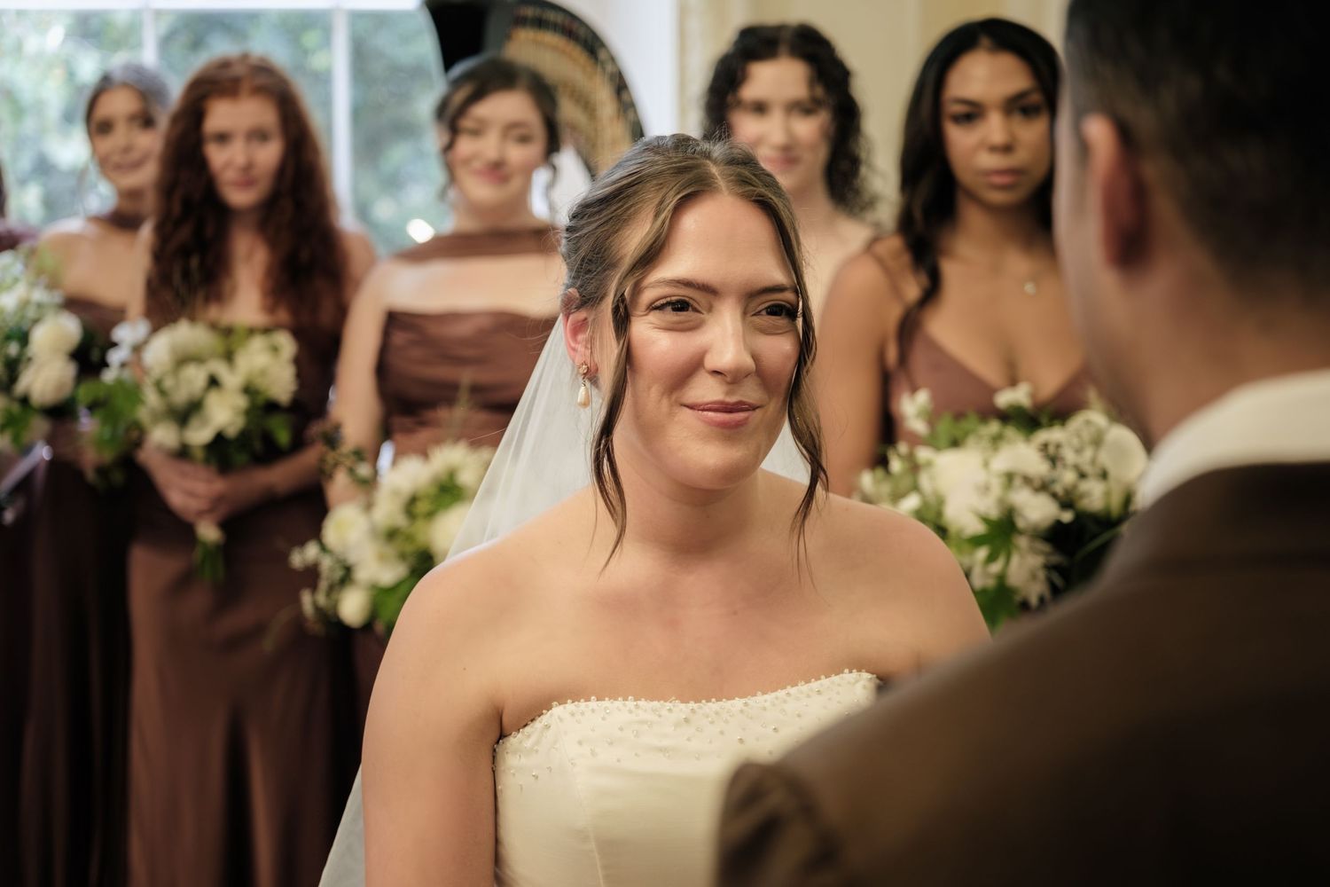 Smiling bride in a white strapless gown and veil faces her groom during the ceremony as bridesmaids in brown dresses hold white bouquets.