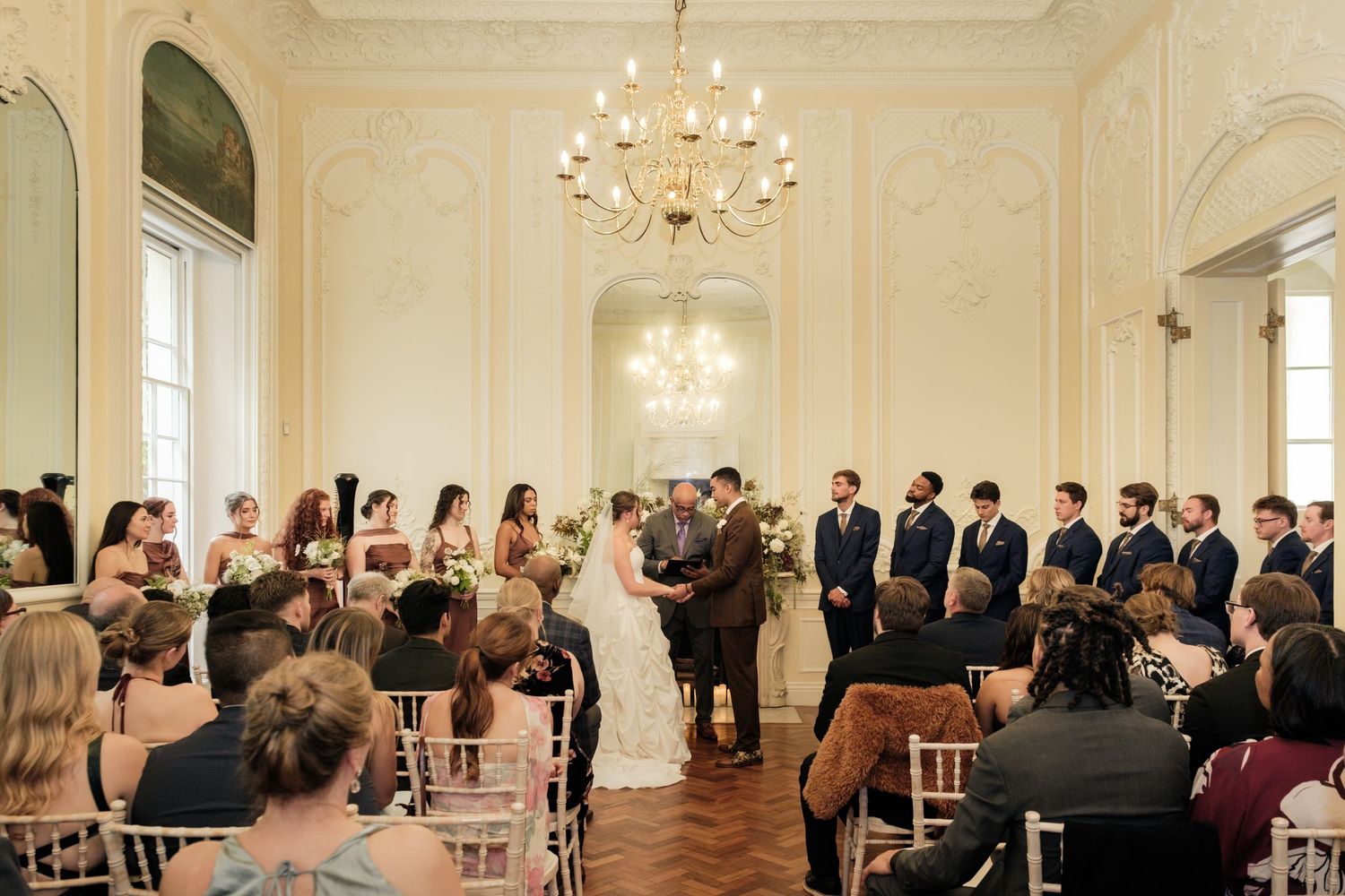 Bride and groom exchange vows before seated wedding guests and groomsmen in an elegant ballroom with a crystal chandelier.