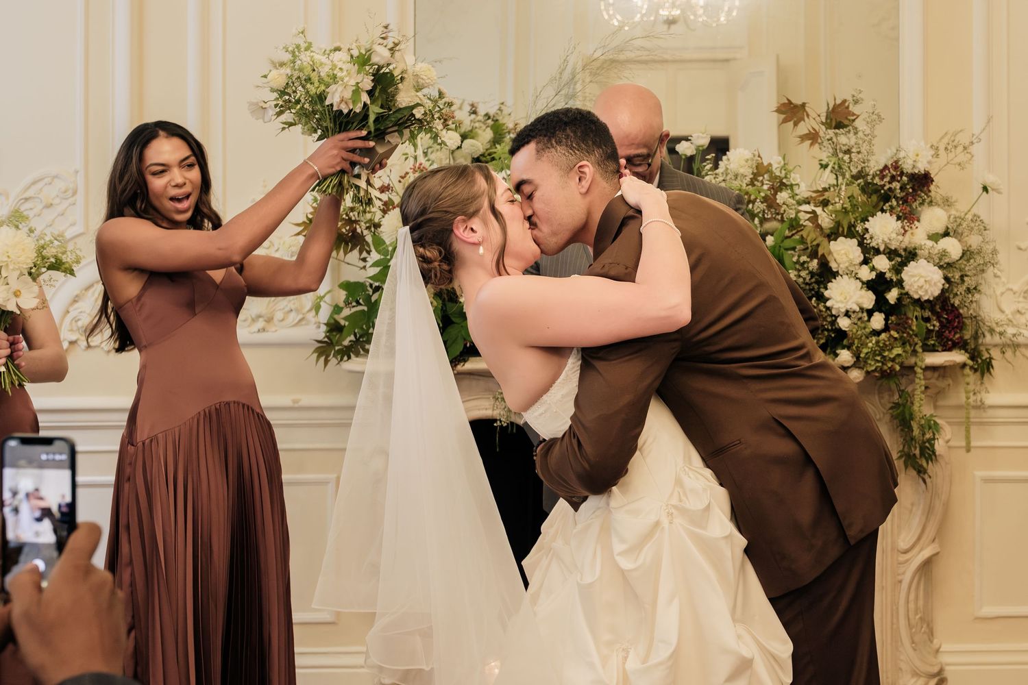 Bride in a white gown and groom in a brown suit share a kiss at the altar as a bridesmaid cheers and a guest films with a phone.