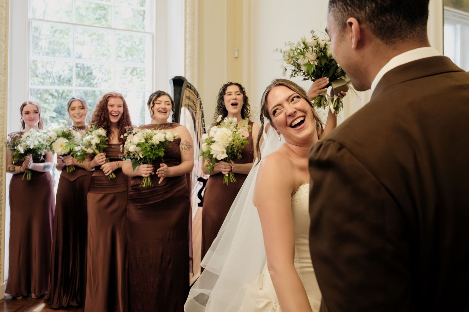 Smiling bride in a white gown and veil laughs with her groom while bridesmaids in brown dresses hold bouquets and smile behind them.