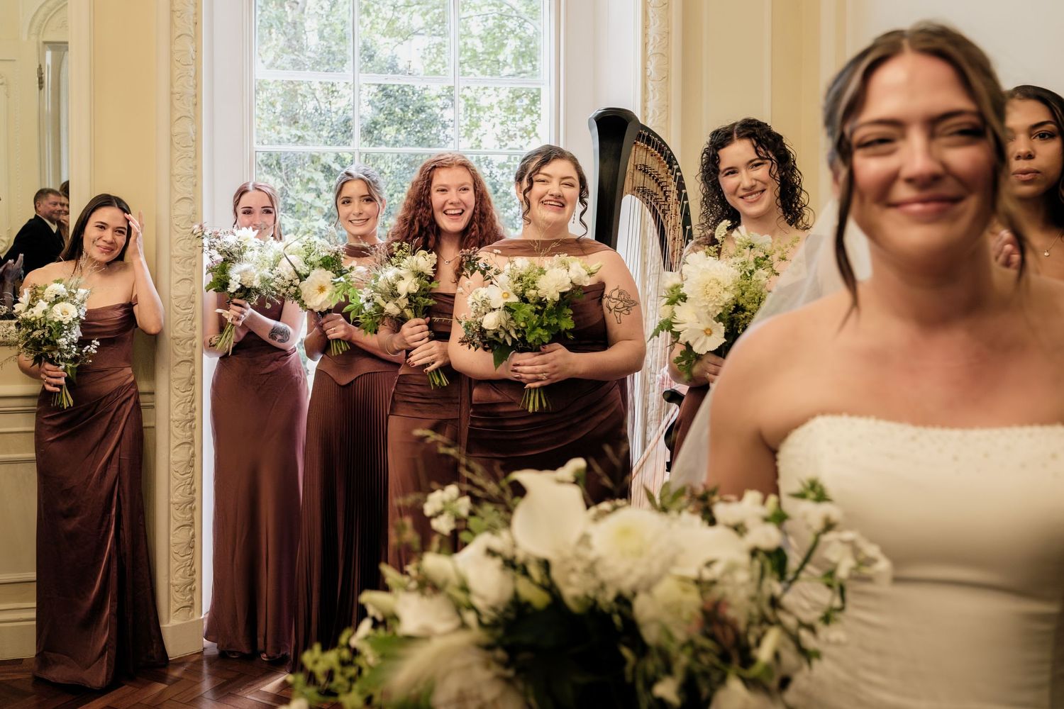 Smiling bride in a white gown holds a white floral bouquet while bridesmaids in brown satin dresses laugh behind her in an elegant room.
