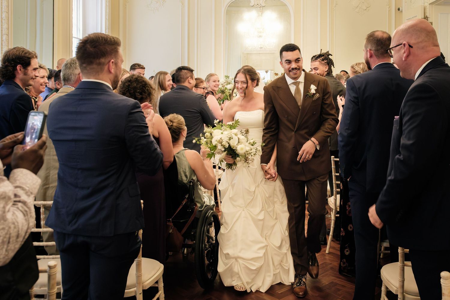 Bride in a white gown holding a bouquet walks down the aisle arm in arm with a groom in a brown suit as wedding guests look on.
