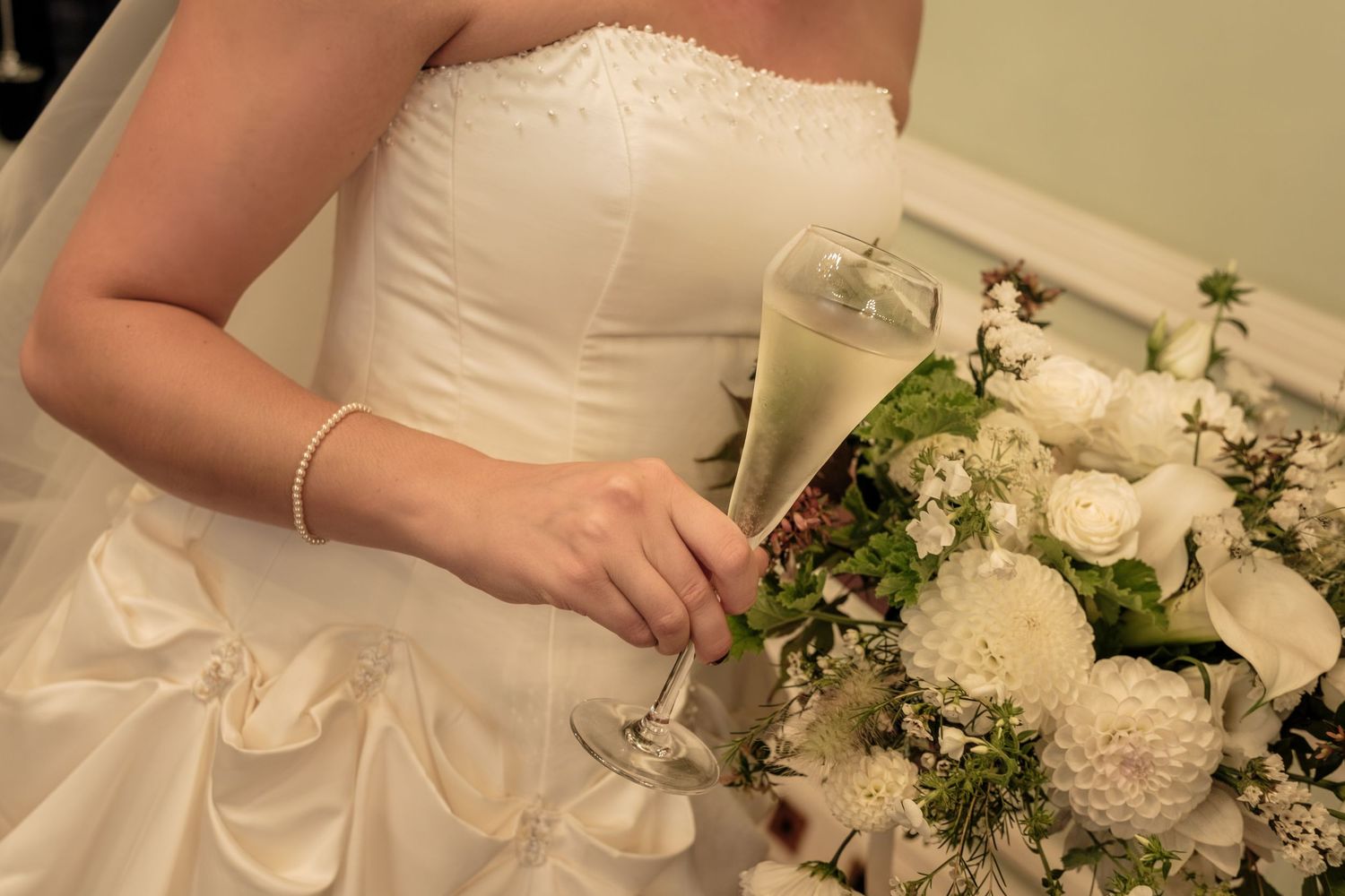 Bride in a strapless white wedding gown holding a glass of champagne and a bouquet of white roses with greenery.