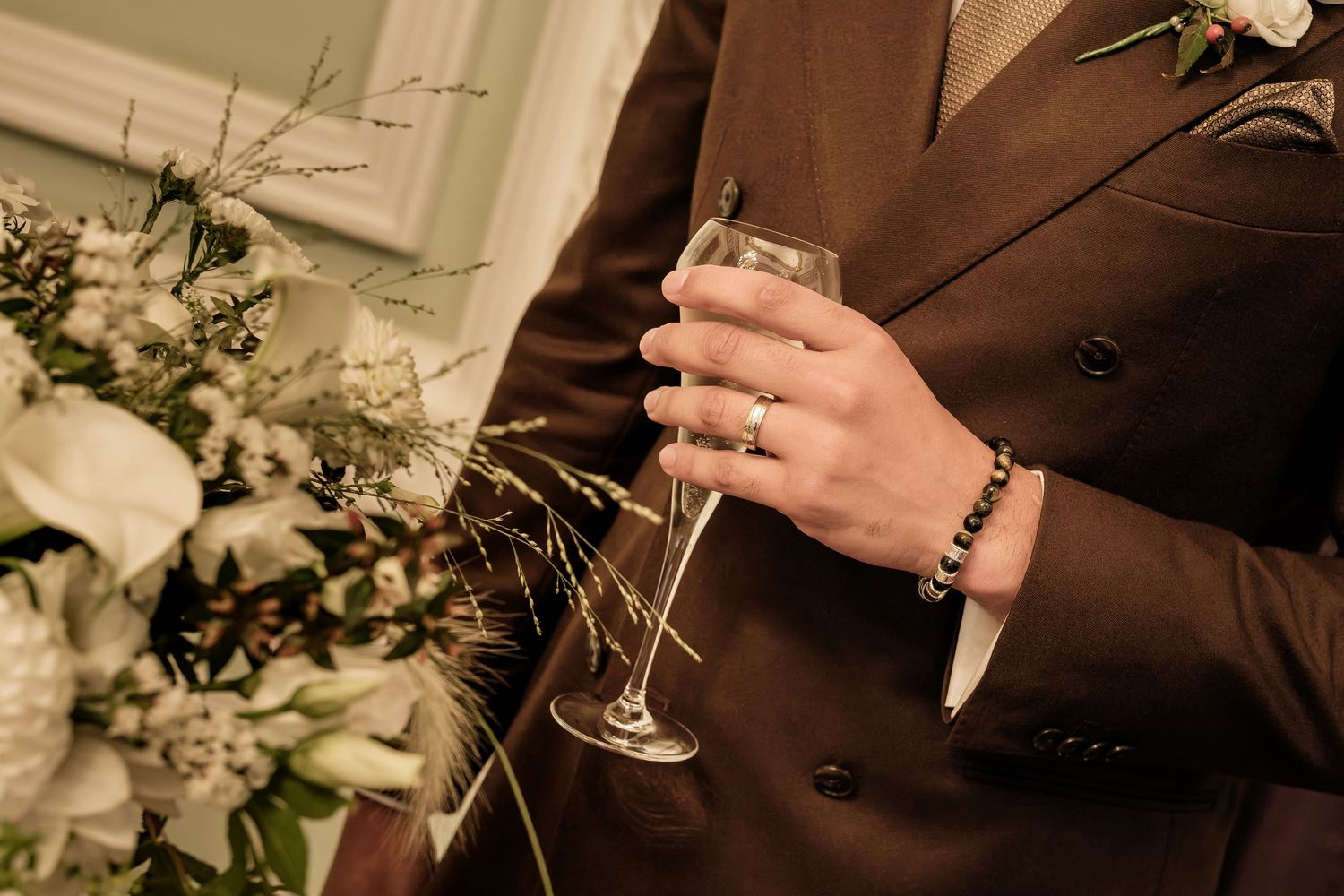 Groom in a brown double-breasted suit holds a champagne flute beside a white floral bouquet at a wedding celebration.