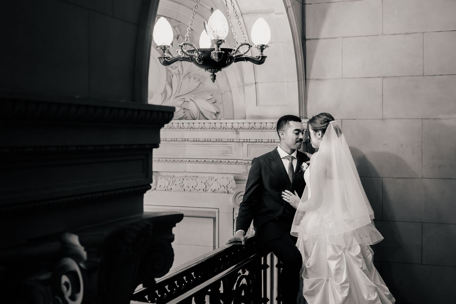 Black and white photo of a bride and groom embracing on an ornate stone staircase beneath a vintage chandelier.