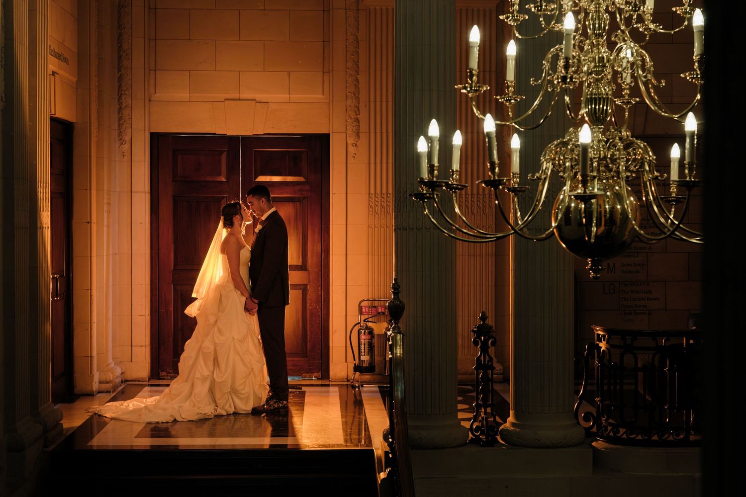 Bride in a white gown and groom in a dark suit embrace in an elegant hall lit by a grand candle chandelier near wooden double doors.