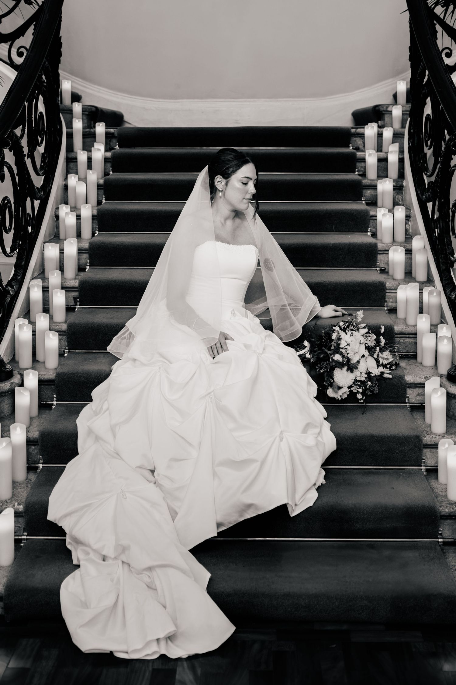 Black and white photo of a bride in a strapless gown and veil sitting on a carpeted staircase lined with candles, holding a bouquet.