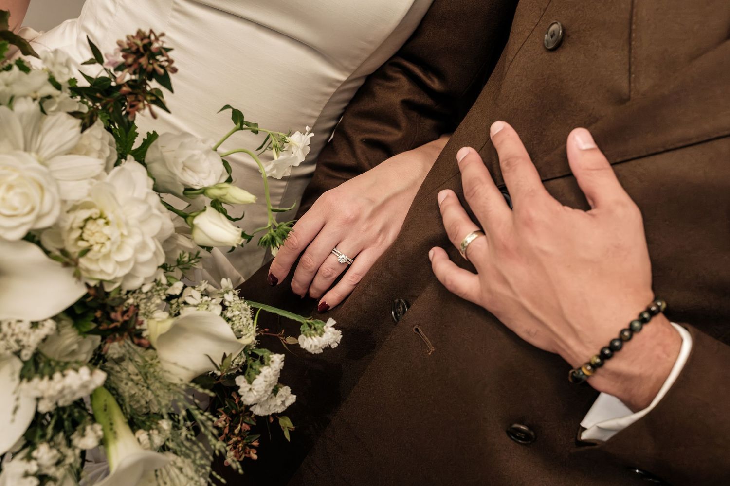 Bride and groom hold hands showing wedding rings, beside a white rose and greenery bouquet, groom in a brown suit with a beaded bracelet.