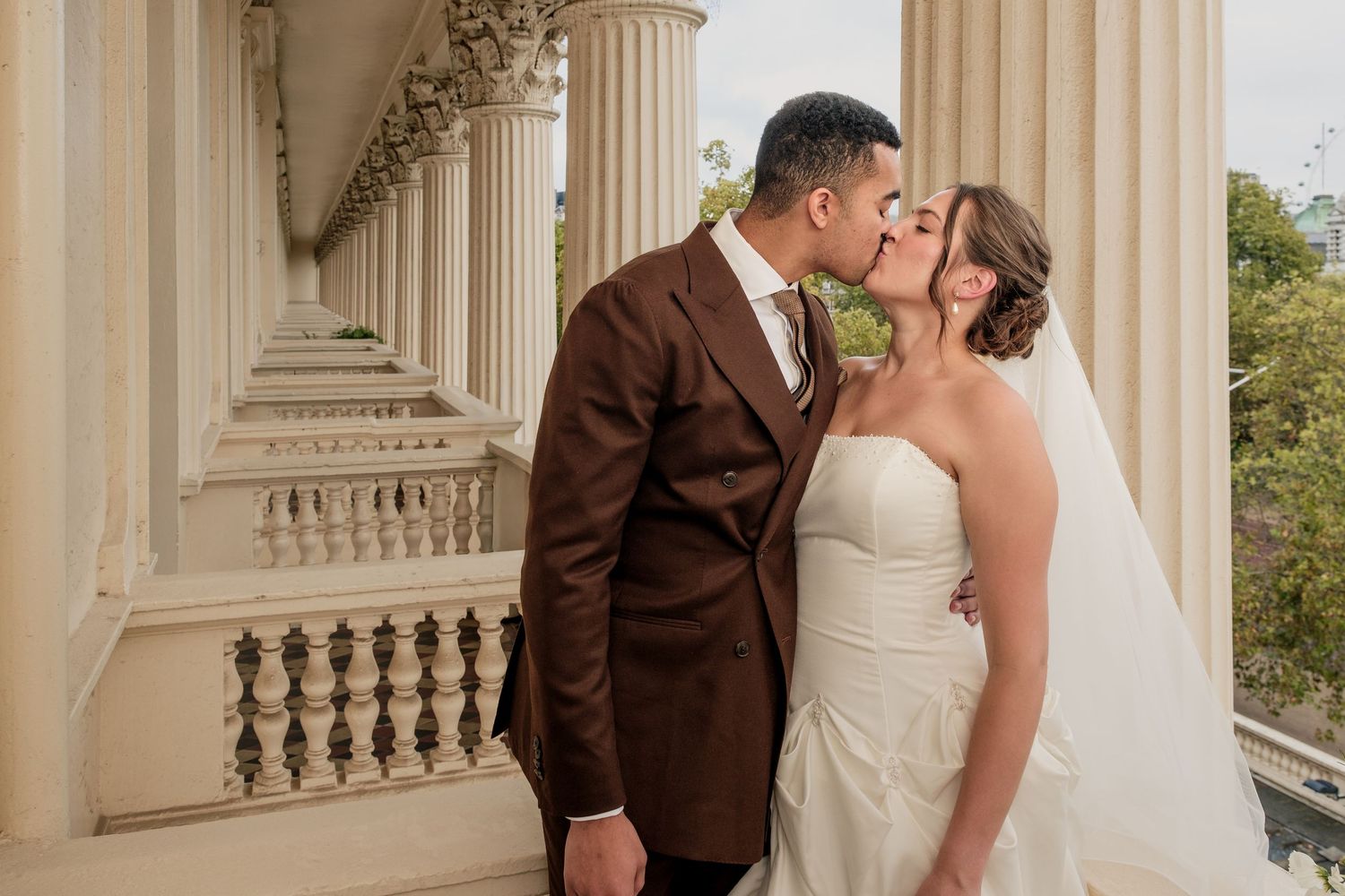 Bride in a strapless white gown and veil kisses groom in a brown suit on a columned stone balcony during their wedding photoshoot.