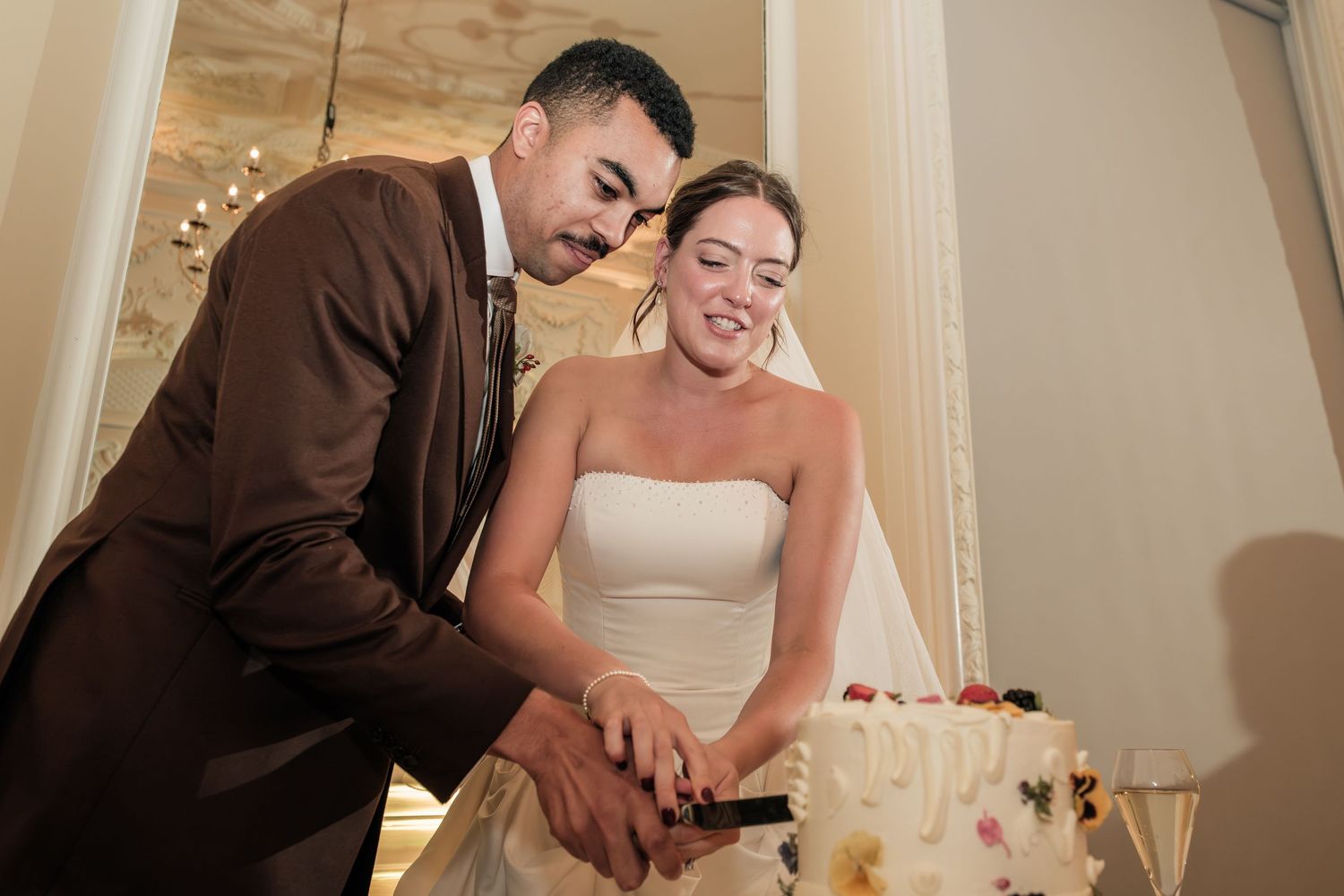 Smiling bride in a strapless white gown and groom in a brown suit cut their floral-decorated wedding cake together at the reception.