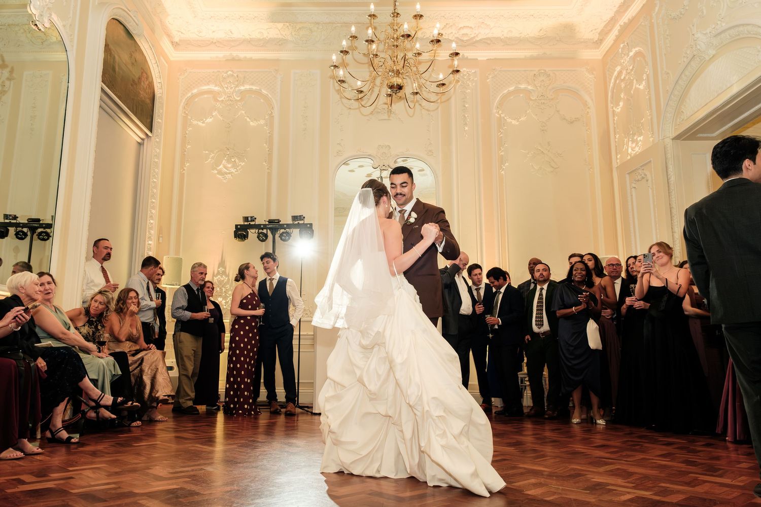 Bride in a white gown and groom in a dark suit share their first dance on a wood floor as guests watch in an elegant ballroom.