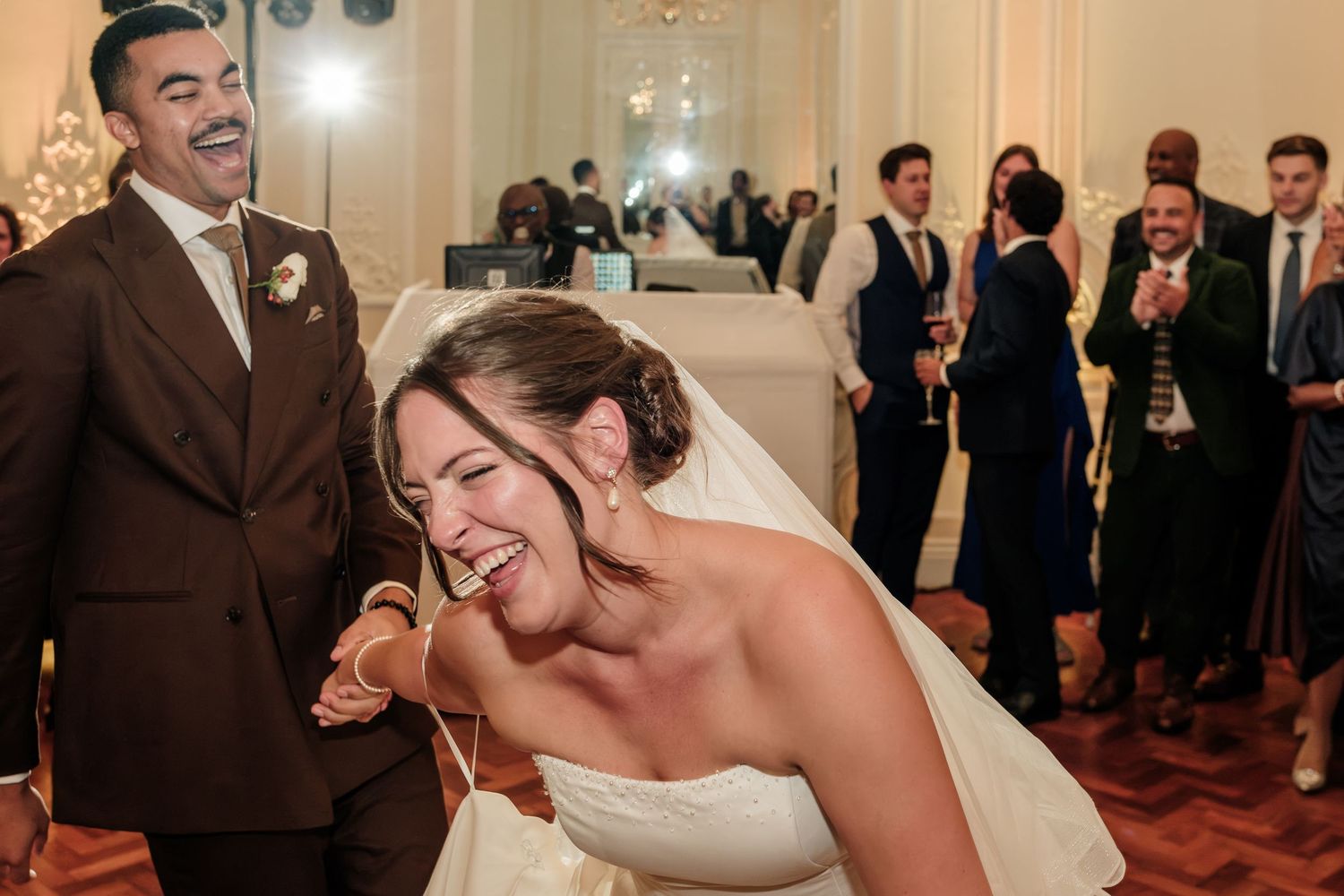 Laughing bride in a white wedding gown leans forward as a smiling groom and guests in formal attire cheer behind her at the reception.