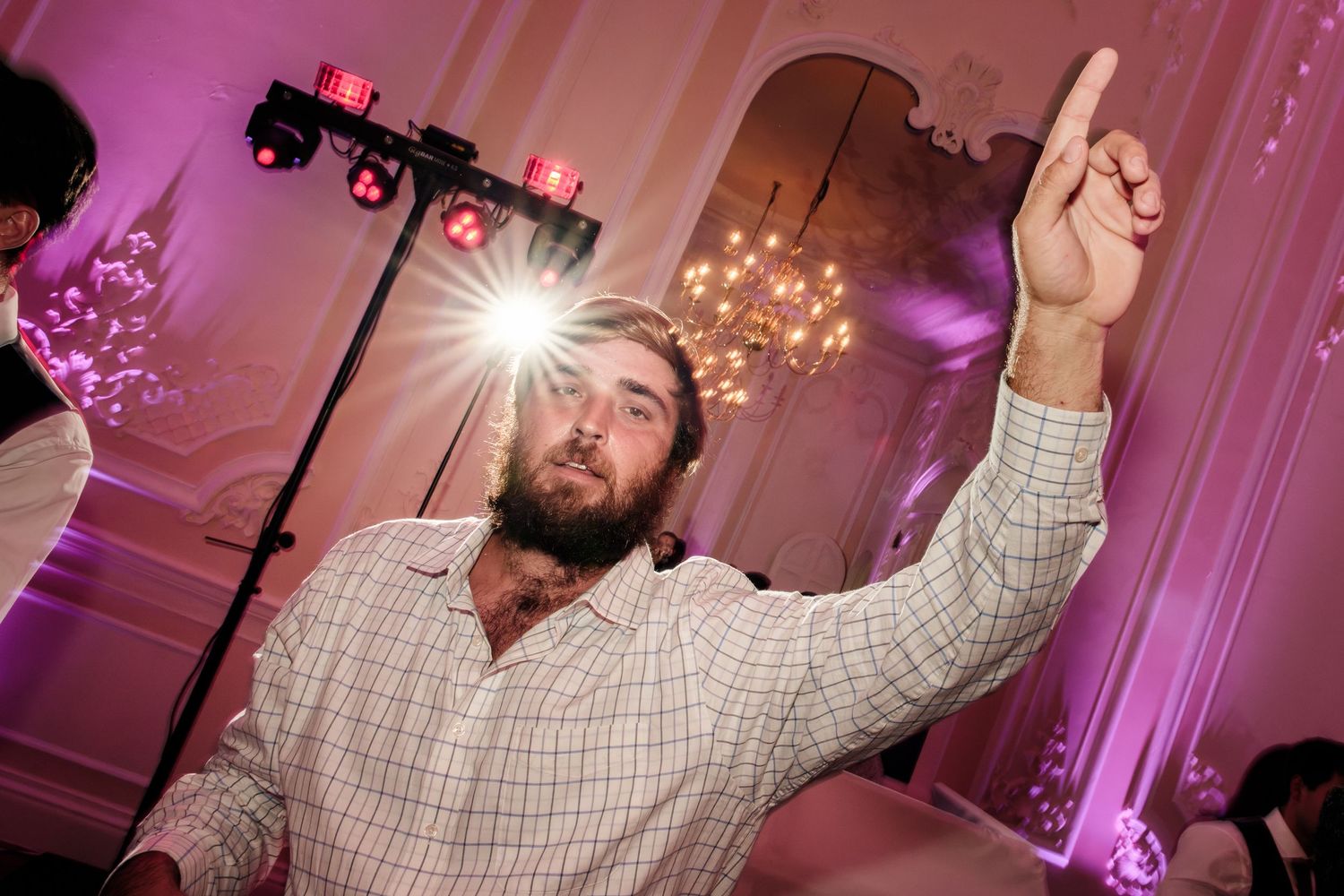 Bearded man in a plaid shirt points upward while dancing at a wedding reception with purple uplighting, stage lights, and a chandelier.