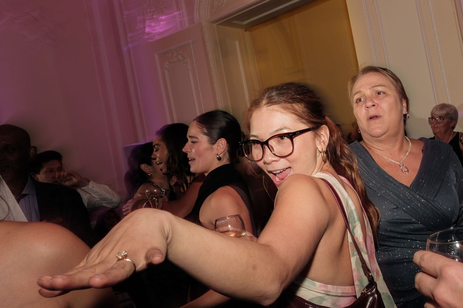 Woman in glasses dances joyfully with her arm outstretched at a lively indoor party while guests laugh and mingle behind her.