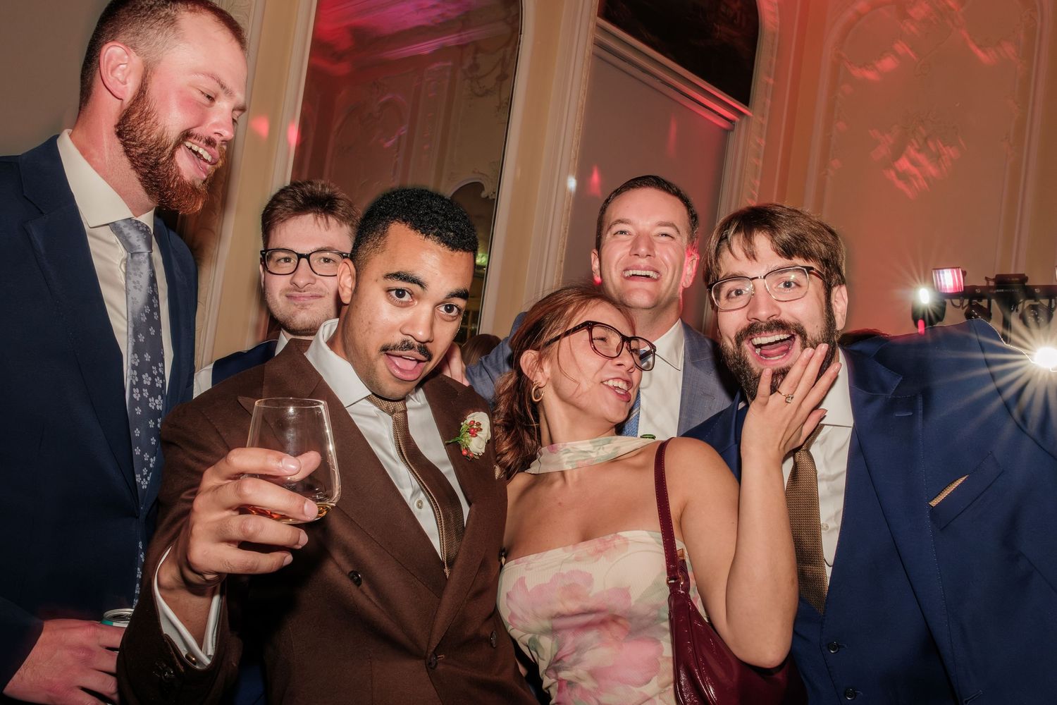 Group of six smiling friends in formal attire posing closely together at an evening party, one man holding a drink in the foreground.