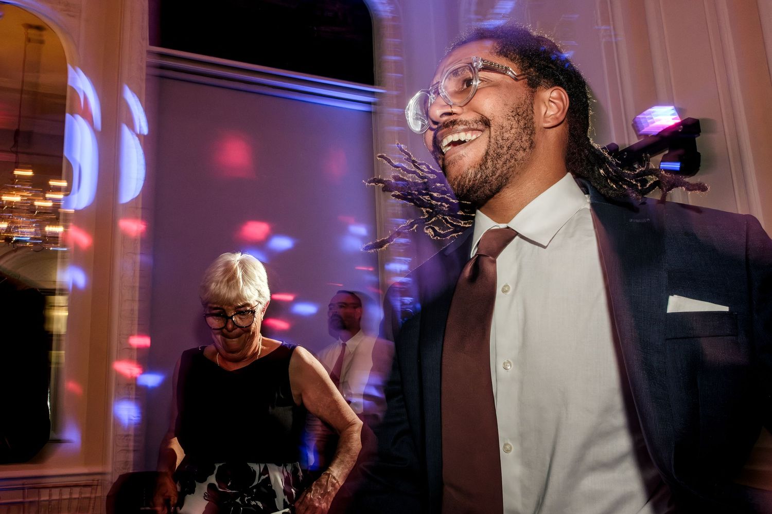 A smiling bearded man in glasses, a dark blazer and a burgundy tie laughs at a lively party while an older woman dances behind him.