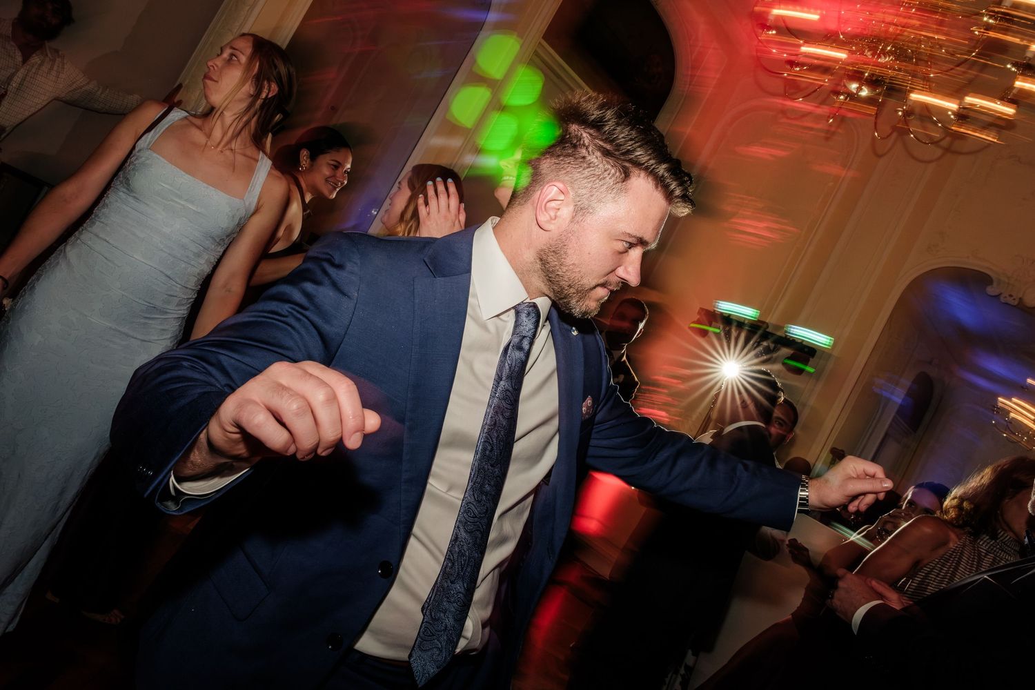 Man in a navy blazer and patterned tie dances energetically at a lively indoor wedding reception with guests and colorful lights behind him.