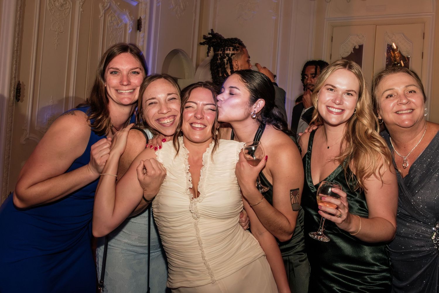 Six smiling women in elegant dresses pose close together at a festive indoor celebration, one kissing another on the cheek.