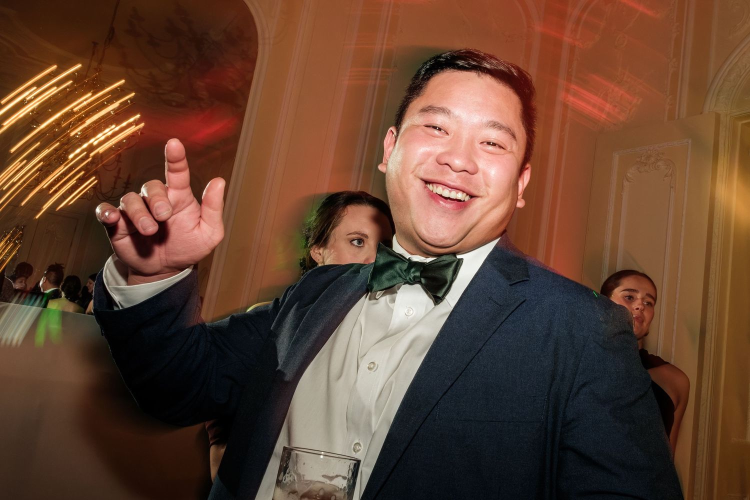 Smiling man in a navy blazer and green bow tie dances at a formal evening party while guests mingle behind him.