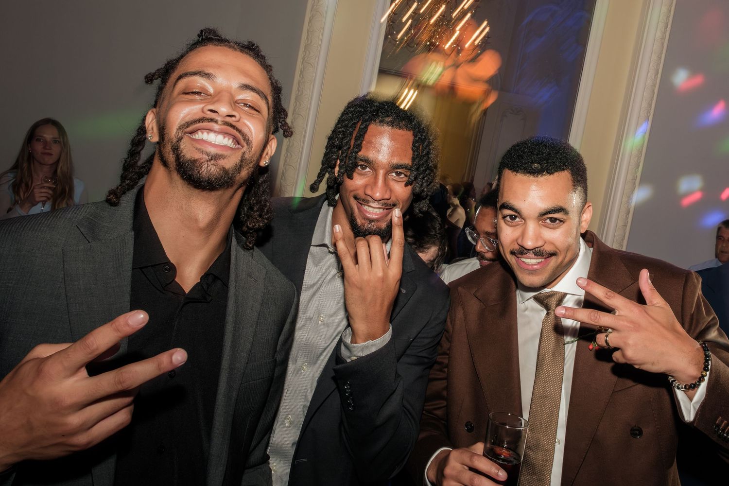 Three smiling men in stylish jackets pose and flash hand gestures at a lively indoor party with warm lighting and guests in the background.