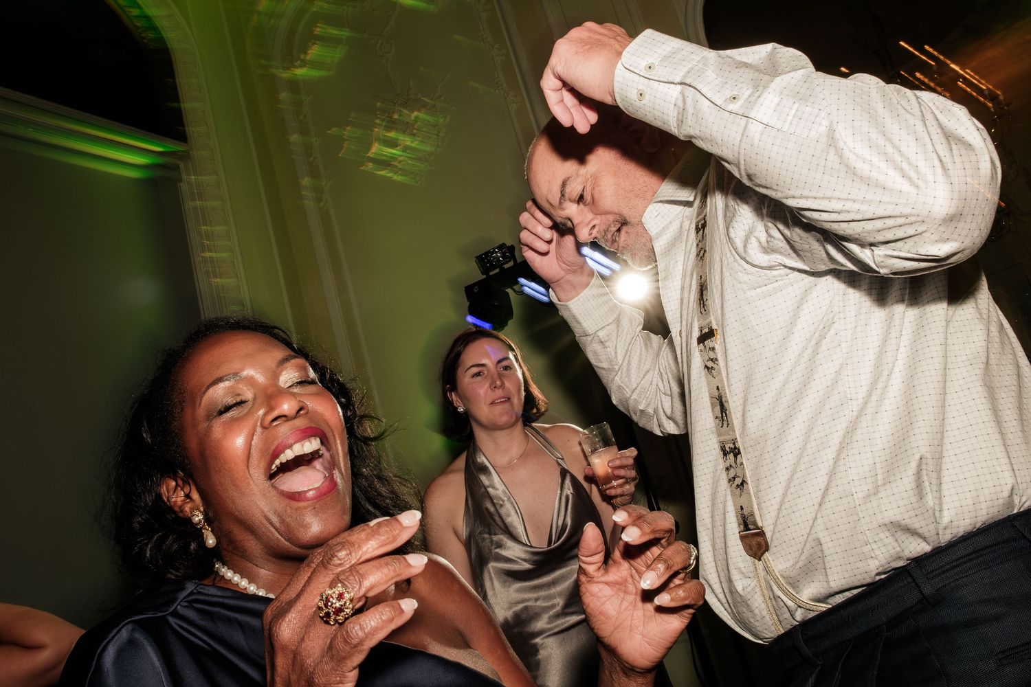 Guests laugh and dance joyfully at a lively party, with a smiling woman in the foreground and a man dancing beside another woman.