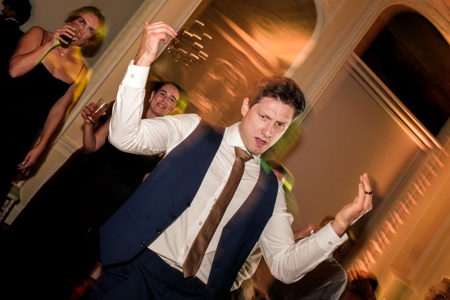 Man in a navy vest and brown tie dances energetically with arms raised at a lively indoor party while guests celebrate behind him.