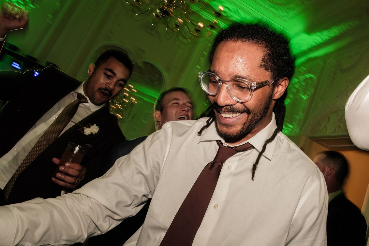 Smiling man with glasses and locs wearing a white dress shirt and brown tie dances at a lively formal party with green lighting.