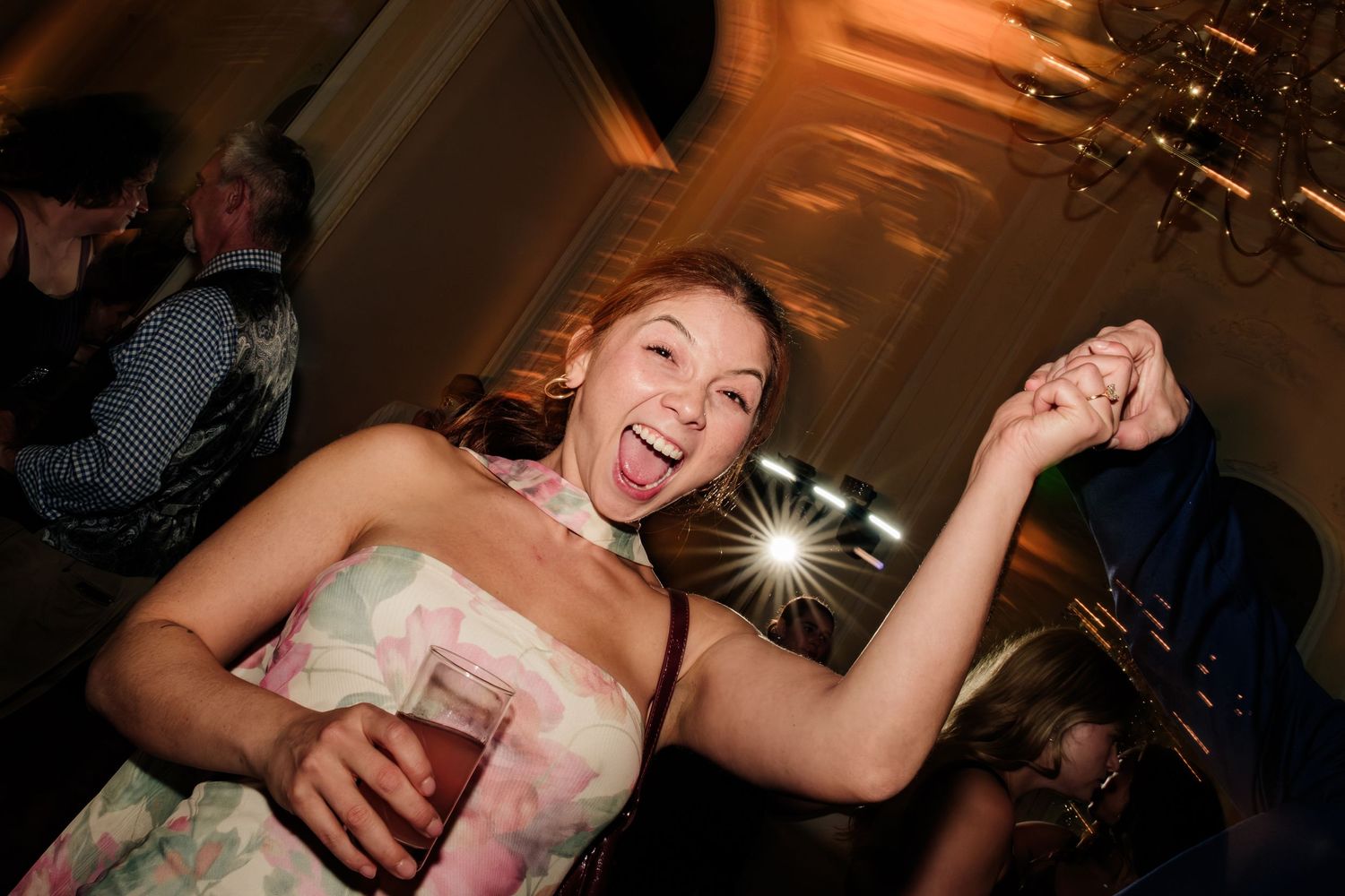 Joyful woman in a floral strapless dress laughs and dances at a party, holding a drink and raising her hand under warm chandelier lighting.